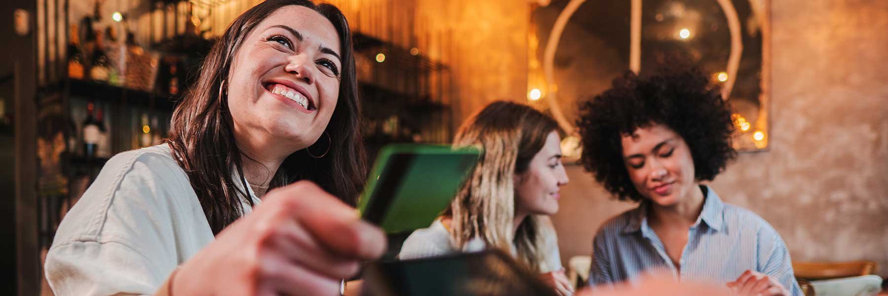 Happy young woman paying bill with a contactless debit card in a restaurant with friends.